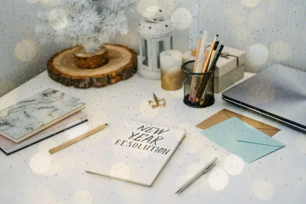 an aerial view of a desk with stationery, notebooks, pens, envelopes and an open notebook with "New Year Resolution" handwritten on. The image has an overlay of sparkles.