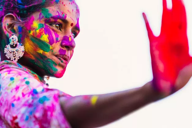 A woman with brown skin and dark hair holds her hand up in a 'stop' motion. She is covered in colourful powder and her hand has red powder on the palm