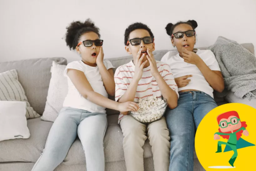 Three young children sat on a sofa with 3D glasses on with popcorn